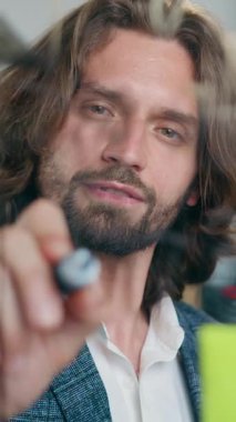 Cam duvarın arkasında duran ve projeyi başlatmak için yeni fikirler üretirken üzerinde önemli işaretler bırakan şık giyimli, deneyimli, yakışıklı, pozitif sakallı adamın portresi.