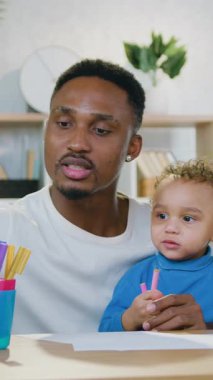 Çağdaş odada, renkli keçeli kalemlerle kağıdın üzerine komik resimler çizerken iki yaşındaki tatlı bebeğiyle ilgilenen 30 yaşında, siyah tenli, çekici bir babanın portresi.