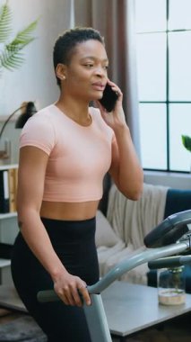 Sağlıklı yaşam tarzı konsepti, spor giyim kuşamında oldukça narin, genç, Afro-Amerikan bayan oyuncunun evde koşu pistinde yürürken cep telefonu konuşmasının keyfini çıkarması.