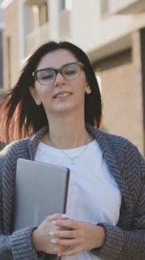 Güzel görünümlü, 30 yaşında, kendine güvenen ve gülümseyen beyaz bir kadının portresi. Şehir caddesinde yürüyen, rüzgarlı bahar havasında dizüstü bilgisayarı tutan bir kadın. Yavaş çekim