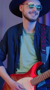 Güzel görünüşlü, gülümseyen, 25 yaşında, siyah şapkalı ve güneş gözlüklü erkek müzisyen gitarla zevkle çalıyor ve konserden önce oyununu geliştiriyor.