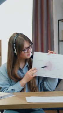 Uzaktan öğrenme konsepti: Çekici, kendine güvenen, kulaklıklı genç bir kız merkez ofiste kameranın önünde oturuyor ve online konferans düzenliyor. Raporları çizelgelerle açıklıyor.