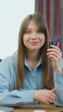 Gülümseyen, kendine güvenen genç bir bayan güzellik blogcusu kozmetik ürünlerini bir elinde tutuyor ve ev stüdyosundaki kameraya el sallıyor.