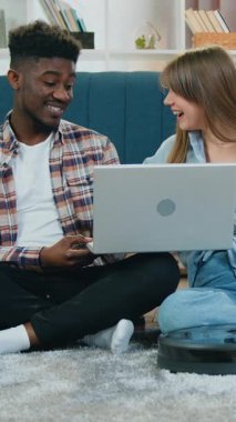 Gülümseyen genç erkek ve kadın çok ırklı çift dizüstü bilgisayar kullanarak modern robot elektrikli süpürgenin yanında pofuduk halı temizliyor..