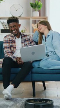 Modern ev konsepti, siyah robot elektrikli süpürge temizlik halısını temizlerken aynı zamanda çekici gülümseyen karışık ırk arkadaşlarının kanepede oturup dizüstü bilgisayarda video izlerken sohbet etmesi.