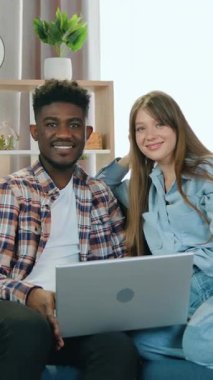 Güzel, gülümseyen genç bir kız, pozitif siyah tenli erkek arkadaşıyla koltukta oturuyor ve mutlu bir yüz ifadesiyle kameraya bakıyor.