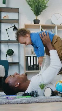 Güzel görünüşlü, şefkatli Afrikalı Amerikalı baba pofuduk halıda rahatlıyor ve kendisini uçakta hissetmesi için küçük oğlunu elleriyle tutuyor.