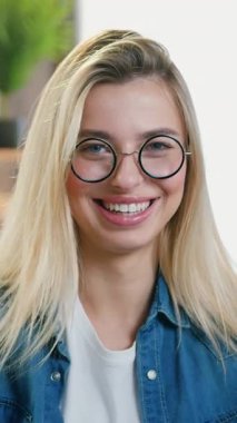 Çok güzel dekore edilmiş oturma odasında kameranın önünde oturan ve samimi bir gülümsemeyle poz veren 25 yaşında, neşeli, açık renk saçlı, gözlüklü bir kızın kadın portresi.