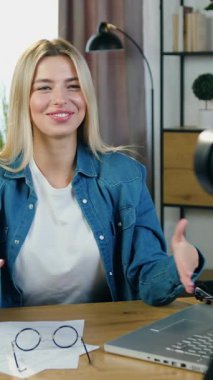 Güzel, neşeli, başarılı, başarılı bir bayan öğretmen stüdyosunda oturuyor ve online takipçileri için canlı yayında sohbet ediyor ve kameraya sembol gösteriyor.