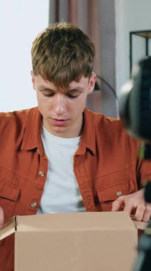 İyi görünümlü pozitif 20-25 yaşındaki erkek blogcu, izleyicileri için karton kutunun paketini açarken video blogunu yayınlarken şaşırdı.