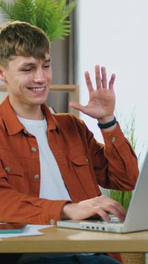 İyi huylu, arkadaş canlısı, 25 yaşında, koyu turuncu tişörtlü, beyaz tişörtlü bir adam, gündüz vakti rahat bir odada masada oturan arkadaşıyla veya akrabasıyla görüntülü konuşma yapıyor.