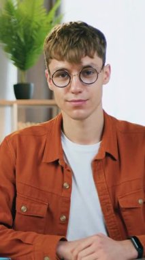 Çekici, pozitif, 25 yaşında, gözlüklü, koyu turuncu gömlekli bir erkek portresi. Çalışma odasında oturmuş, kameraya sembol ile bakıyor.