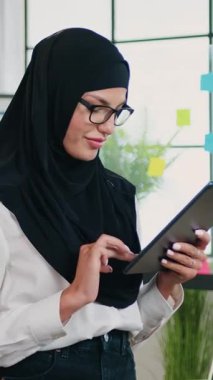 Tatmin edici deneyimli, 30 yaşında, gözlüklü, siyah tesettürlü başarılı Arap iş kadını güzel dekore edilmiş çalışma alanında tablet üzerinde çalışıyor.