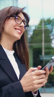 Takım elbiseli başarılı bir beyaz iş kadını, bir bankta oturuyor, cep telefonuyla mesajlaşırken gülümsüyor. Arka planda şehir binaları ve gökyüzü var..