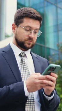 Güneşli bir yaz gününde ceketli ve kravatlı beyaz bir adam akıllı telefon kullanıyor ve arkasında modern cam bir bina bulunan bir mesaj yazıyor.