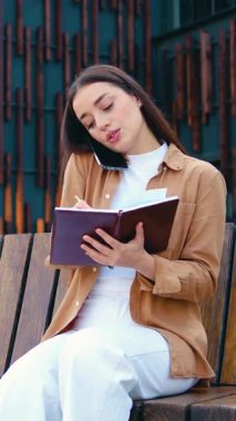 Gülümseyen serbest çalışan bir kadın cep telefonuyla konuşuyor ve dışarıda modern ofis binası hakkında bankta otururken uzaktan not alıyor.