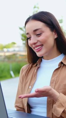 Genç bir kız çevrimiçi video görüşmesi sırasında gülümsüyor güneşli bir günde laptopuyla dışarıda oturuyor ve temiz havanın tadını çıkarıyor.
