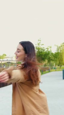 Neşeli genç bir kadın günlük kıyafetleriyle şehir parkında geziniyor, kameraya neşe içinde poz veriyor, açık hava ortamında neşeli ve kaygısız bir anı yakalıyor.