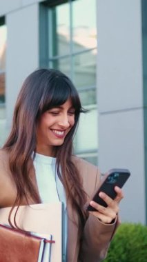 Genç, beyaz bir kadın güvenli bir şekilde modern cam ofis binasının yanında yürüyor, elinde bir dizüstü bilgisayar ve bir defter, akıllı telefonunu kontrol ederken gülümsüyor..