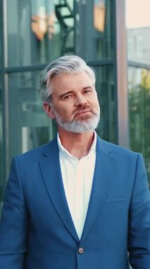 Şehrin dışında duran, kolları çaprazlanmış kameraya güvenle gülümseyen, arka planda modern cam ofis binası, açık havada duran kıdemli sakallı iş adamının portresi.