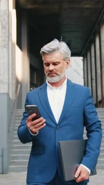 Kendine güvenen Kafkasyalı iş adamı resmi giyinmiş, akıllı telefon kullanıyor, modern ofis binasının önünde yürürken mesaj yazıyor..