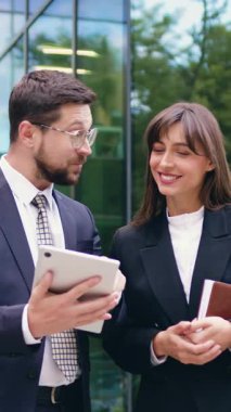 Elinde tablet olan gülümseyen bir iş adamı ve elinde not defteri olan bir iş kadını modern bir ofis binasının önünde durup başarılı bir projeyi kutlamak için birbirlerine beşlik çakıyor.