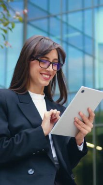 Resmi giyinmiş genç bir iş kadını modern bir ofis binasının önündeki bankta oturuyor, tablet üzerinde çalışıyor ve bir müşteriye yapay zeka sohbet robotu kullanarak bir mesaj yazıyor.