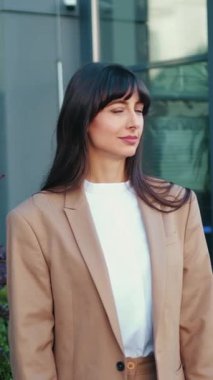 Genç ve gülümseyen bir iş kadını, kameraya poz veriyor. Kollarını vücudunun önünde kavuşturuyor. Arka planda modern bir ticari bina var..