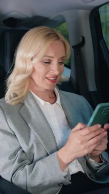 Arabasının arka koltuğunda akıllı telefonu olan resmi giysili beyaz bir kadın. Orta yaşlı bir kadın arabada cep telefonu kullanır. İnternet ve sosyal medya. Yaşam tarzı. Yavaşlama Hareketi