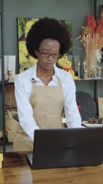 Dizüstü bilgisayarda çalışan sevimli, ciddi, siyah tenli bir kadın işçi. Rafların yanında duruyor. Önü güzel hatıralarla dolu.