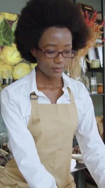 Güzel görünümlü, kendinden emin, 25 yaşında, neşeli, beyaz önlüklü, el yapımı hediyelik eşya dükkanında çalışırken kameraya gülümseyen Afro-Amerikan kızı.