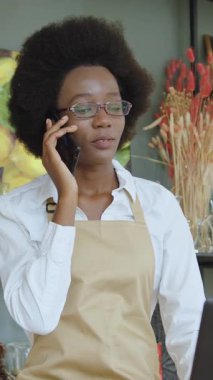 Çekici, kendine güvenen, gözlüklü, farklı hediyelik eşya ve dekorlarla rafların yanında duran ve hoş bir telefon konuşması yapan Afro-Amerikan kadın satıcı.