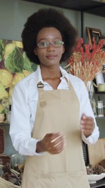 İyi görünümlü, yüksek ruhlu, siyah tenli genç bir kadın. Kameraya bakıyor, kollarını kavuşturup, güzel hediyelik eşya dükkanına bakıyor.