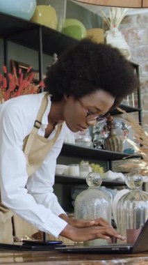 Mükemmel yoğunlaşmış 25-30 yaşlarındaki siyah derili kadın hediyelik eşya dükkanları görevlisi farklı şeylerin fotoğraflarını çekiyor ve bilgisayarına yavaş çekim internet sitesi için fotoğraflar yüklüyor.