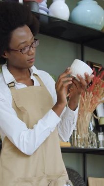 Güzel görünümlü, 25 yaşında, bej önlüklü, el yapımı hediye dükkanında çalışan, elinde beyaz bardak ve şarap kadehiyle dövülmüş mü yoksa kırılmış mı diye bakan Afro-Amerikan kadın bir satıcı.