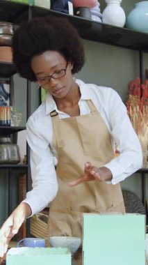 Güzel, çalışkan, siyah tenli, Afrikalı saç stili bir bayan satıcı. Hediye kutularına beyaz bardak ve şarap koyarken el yapımı hediyelik eşya dükkanında çalışıyor.