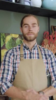 Güzel görünüşlü, pozitif sakallı, sıradan kıyafetler ve bej önlüklü, rahat bir hediyelik eşya dükkanında çalışırken veya hediye dükkanında çalışırken kameranın önünde duran 30 yaşında bir satıcı.