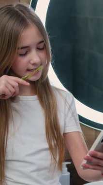 Dişlerini fırçalayan ve akıllı telefon kullanarak tuvaletteki sosyal ağları kontrol eden sevimli küçük bir kız. Elinde cep telefonu olan ve mesaj yazan bir kız..