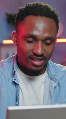 Keyifli, neşeli, kendinden emin Afrikalı Amerikalı arkadaşlarıyla bilgisayar başında renkli gece ışıklarıyla güzel dekore edilmiş ev ofisinde sohbet ediyor.