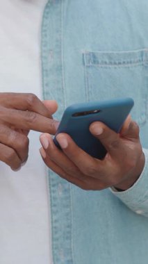 Beyaz tişörtlü ve açık mavi tişörtlü, tanınmayan modern Afro-Amerikan vatandaşı gündüz vakti sokak kafesinin yanındaki akıllı telefonunu kullanıyor, 4K