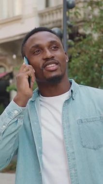 Neşeli, neşeli, neşeli, 25 yaşında, modaya uygun kıyafetleri olan, modaya uygun giyinmiş Afro-Amerikan. Eski şehirdeki bir kafede arka planda akıllı telefondan konuşan, gündüz vakti, 4k.