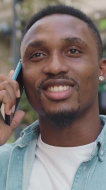 İyi huylu, 25-30 yaşlarında, modern stil sahibi, modaya uygun sakallı ve küpeli Afro-Amerikan 'ın ön görüntüsü. Arka plandaki açık hava kafeteryasında arkadaşlarıyla cep telefonundan konuşan 4k