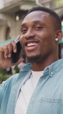 Sevimli bir gülümseme 30 yaşında, modaya uygun sakallı Afro-Amerikan açık hava kafesinin arka planında açık mavi gömlekli, akıllı telefondan konuşan, ağır çekim, 4k