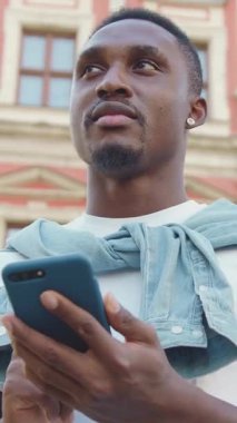 İyi görünümlü, genç, Afro-Amerikan, havalı sakallı, yuvarlak küpeli şehir mimarisi geçmişindeki arkadaşına mesaj yazarken etrafa bakınıyor.