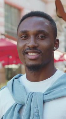 Zarif sakallı, küçük yuvarlak küpeli, modern açık hava kafelerinin yakınındaki kameraya bakan iyi huylu bir adamın portresi.