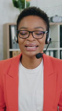 Çekici gülümseme deneyimli Afroamerikan kadın ofis yöneticisi kablosuz kulaklıkla dinleyiciler için online konferans düzenlerken kendi kabinesinde web kamerasının önünde oturuyor.