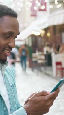 Güzel görünümlü, mutlu, 30 yaşında, modern Afrikalı Amerikalı 'nın portresi. Kulaklarında yuvarlak küpeler var. Güneşli bir günde rahat sokak kafelerinin arka planında mobil uygulamalar var.