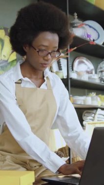 İyi görünümlü, kasıtlı, 25 yaşında, stil sahibi Amerikalı bir kızın portresi. Bilgisayarın önünde duruyor ve hediye dükkanında farklı güzel şeylerin arka planı üzerinde çalışıyor.