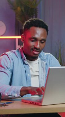 İyi görünümlü, neşeli, modern, 30 yaşında, siyah tenli erkek işçi akşamları iş yerinde oturuyor ve dizüstü bilgisayardaki işini başarıyla bitirmekten zevk alıyor.