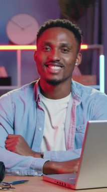İyi görünümlü, yüksek yetenekli, neşeli, siyah tenli bir adam. Ev ofisinin rahat masasında oturuyor. Renkli gece ışıklarıyla ve dizüstü bilgisayarındaki işi sırasında kameraya bakıyor.